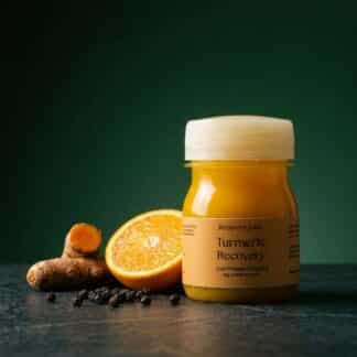 Recovery Juice Turmeric Recovery shot on dark green with fresh turmeric root, orange and black peppercorns